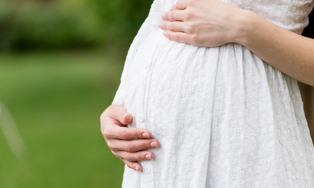 partial view of pregnant woman in white dress touching belly while standing in park