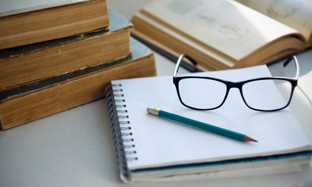 Eldre bøker ligger på et bord med en notatblokk og briller